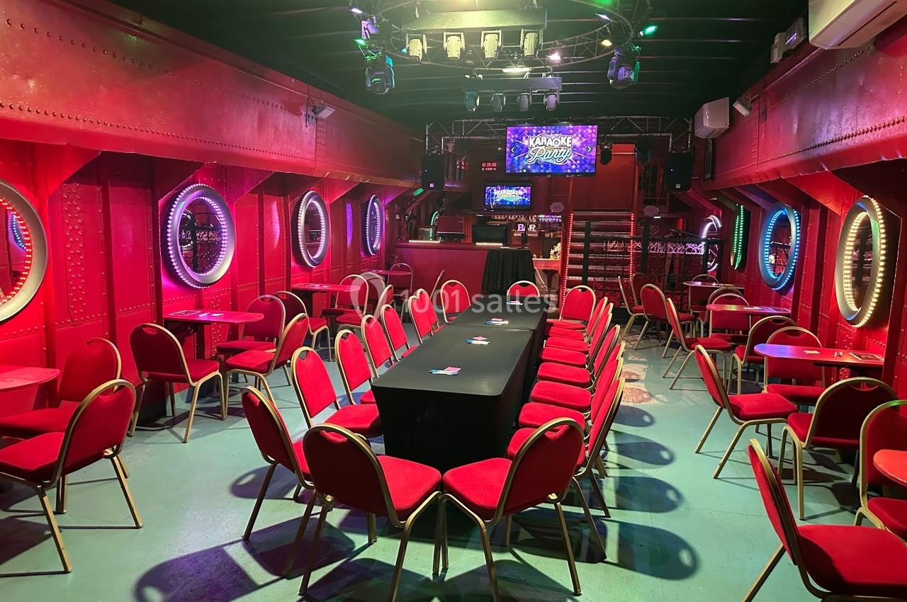 Salle avec des murs rouges, des hublots lumineux, des chaises rouges alignées autour d'une table noire et une scène au fond.