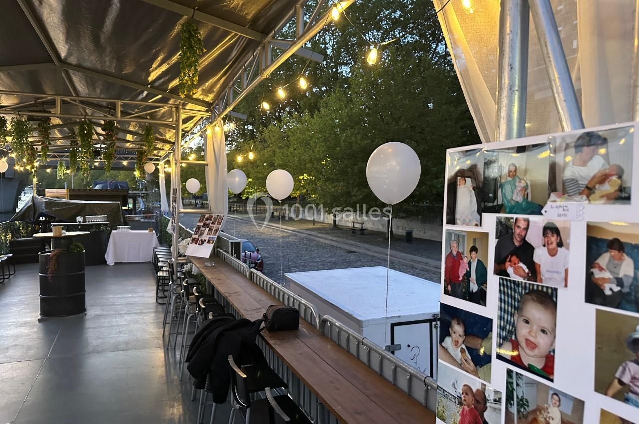 Terrasse décorée avec guirlandes lumineuses, ballons blancs et photos, donnant sur une promenade arborée au bord de l'eau.