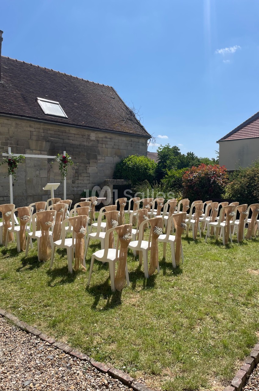 Chaises blanches décorées disposées en rangées sur une pelouse, face à une arche fleurie dans un jardin ensoleillé.