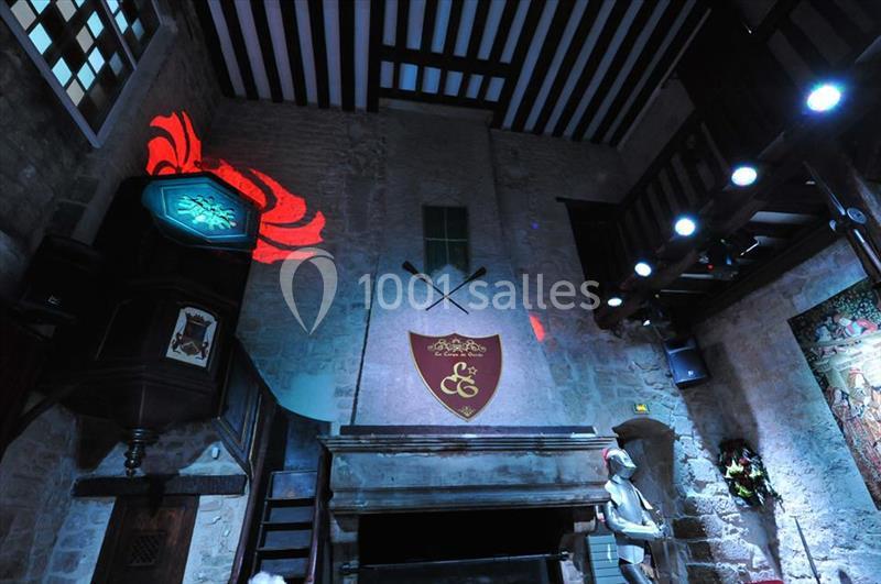 Salle médiévale avec cheminée, blason rouge, armure et éclairages colorés projetant des motifs sur les murs en pierre.
