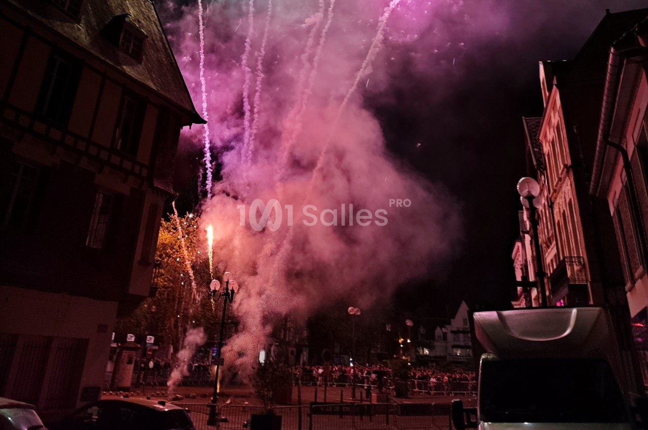 Feux d'artifice illuminant une place urbaine la nuit, avec des bâtiments et une foule en arrière-plan.
