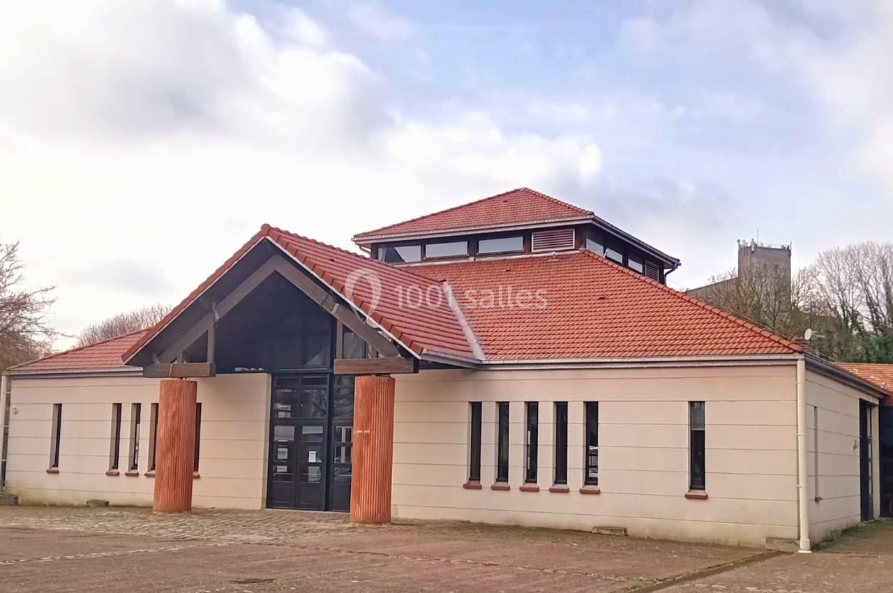 Bâtiment moderne à toit rouge avec une entrée en bois, entouré d'arbres et d'un ciel partiellement nuageux.