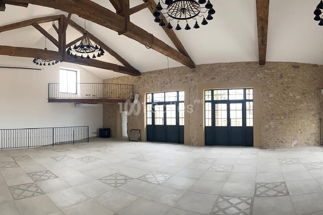 Salle spacieuse avec poutres en bois apparentes, murs en pierre, grandes fenêtres et mezzanine métallique.