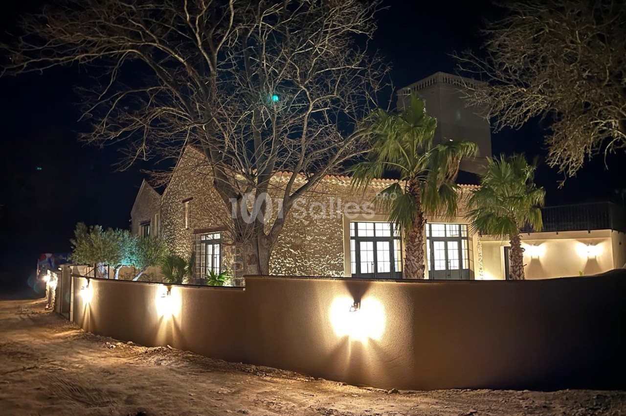 Maison en pierre éclairée la nuit, entourée d'un mur avec des lampes murales et des arbres dénudés.