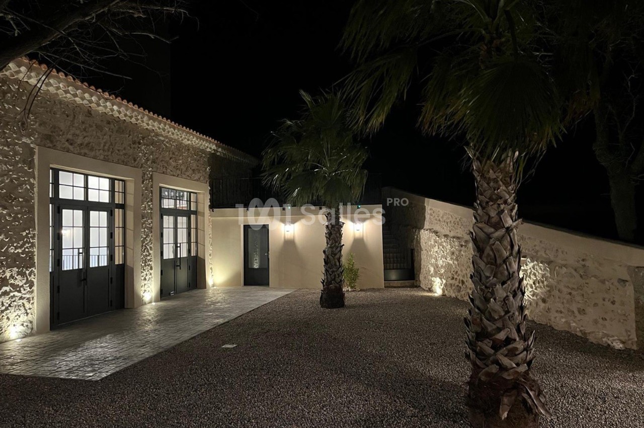 Cour extérieure éclairée de nuit, avec façade en pierre, grandes portes vitrées et palmiers sur sol gravillonné.