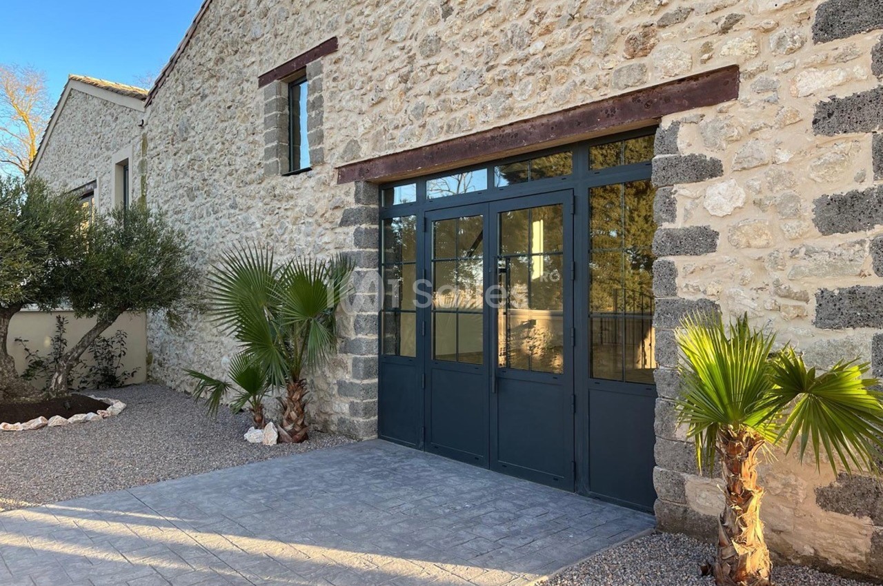 Façade en pierre avec une grande porte vitrée noire, entourée de palmiers et d'un sol en gravier.