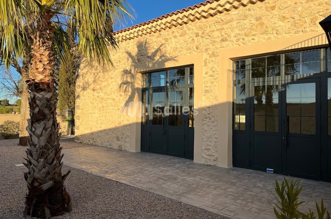 Façade en pierre d'un bâtiment avec grandes portes vitrées, éclairée par le soleil, entourée de végétation.