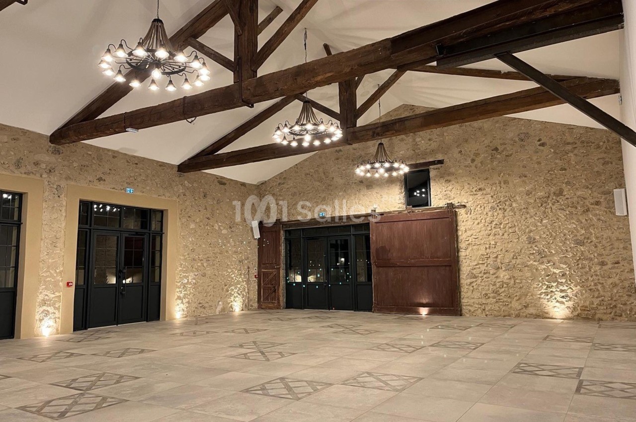 Grande salle avec murs en pierre apparente, poutres en bois au plafond, sols carrelés et éclairage suspendu.