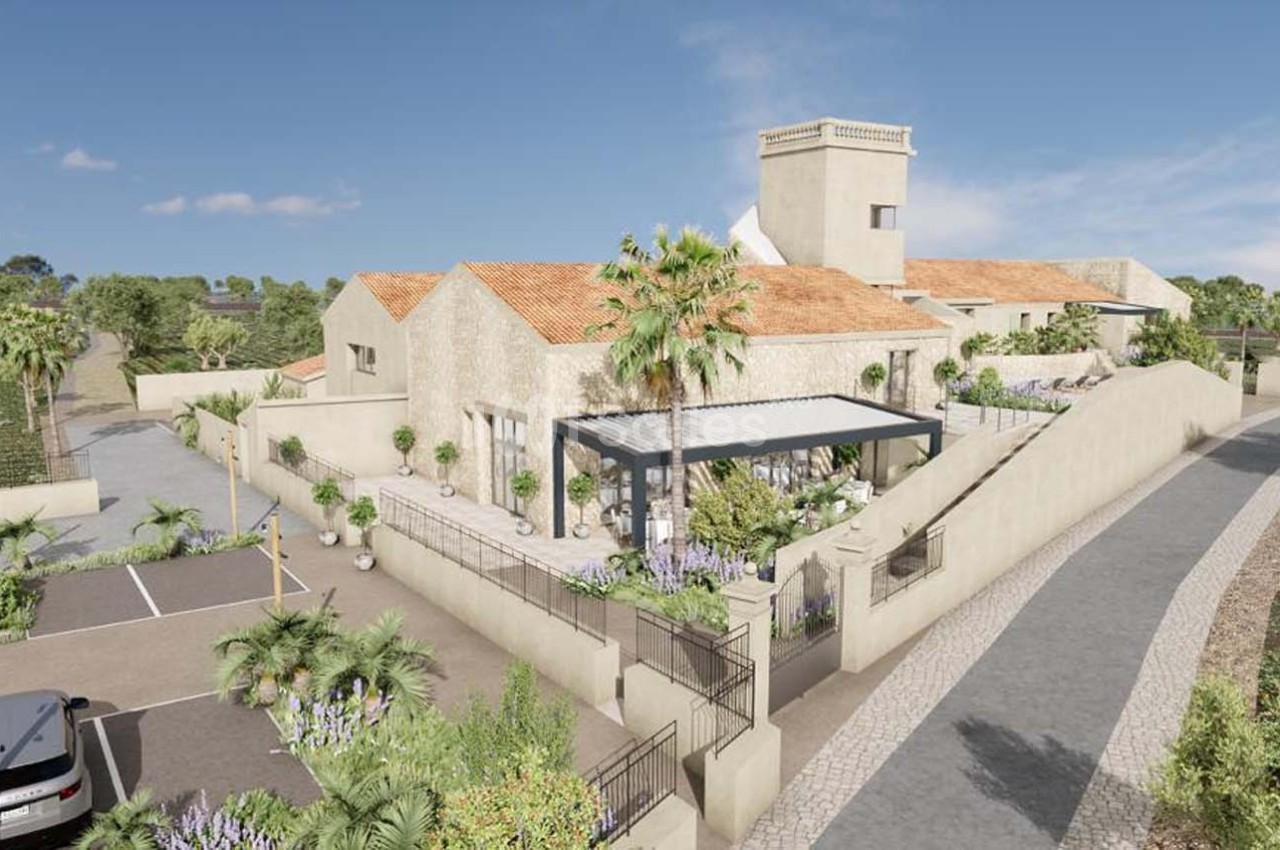Vue d'un bâtiment en pierre avec toit en tuiles, entouré de végétation, d'une terrasse et d'un parking adjacent.