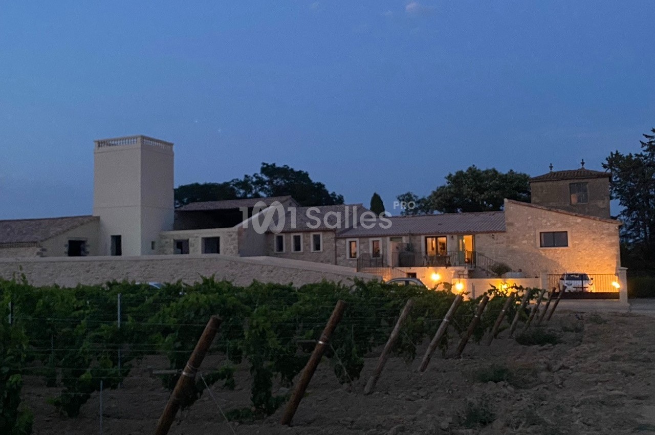 Bâtiment en pierre éclairé à la tombée de la nuit, entouré de vignes et d'arbres.