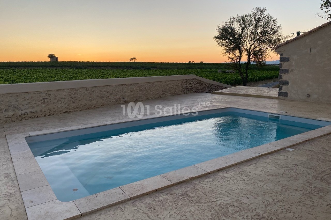Piscine extérieure entourée de dalles en pierre, avec vue sur un champ et un coucher de soleil à l'horizon.