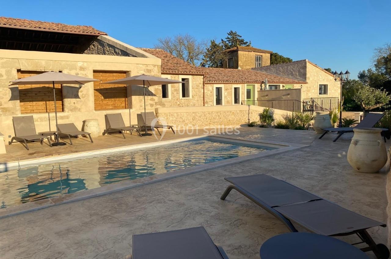 Terrasse en pierre avec piscine, chaises longues, parasols et bâtiments en pierre sous un ciel dégagé.