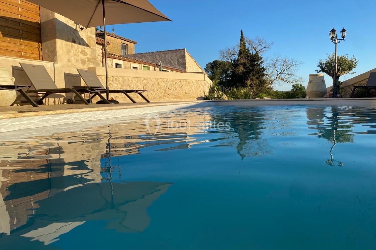 Piscine extérieure calme entourée de chaises longues, parasols et bâtiments en pierre sous un ciel dégagé.