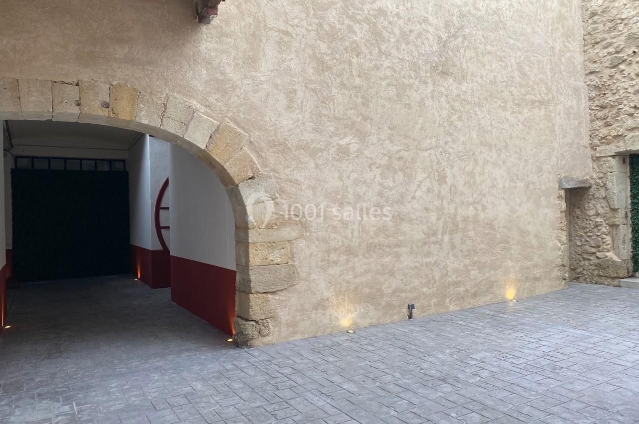 Cour intérieure avec sol pavé, mur en pierre beige, arche en pierre et ouverture vers un passage sombre.