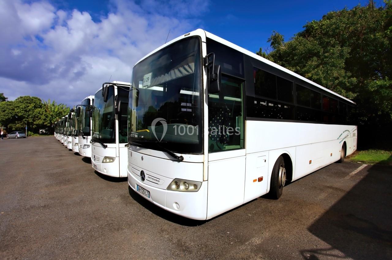 Alignement de plusieurs bus blancs stationnés sur un parking, entourés de végétation et sous un ciel partiellement nuageux.