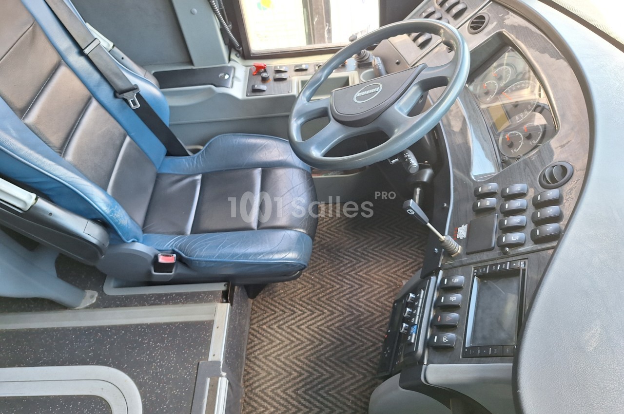 Poste de conduite d'un bus avec siège en cuir bleu, volant, tableau de bord et commandes visibles.