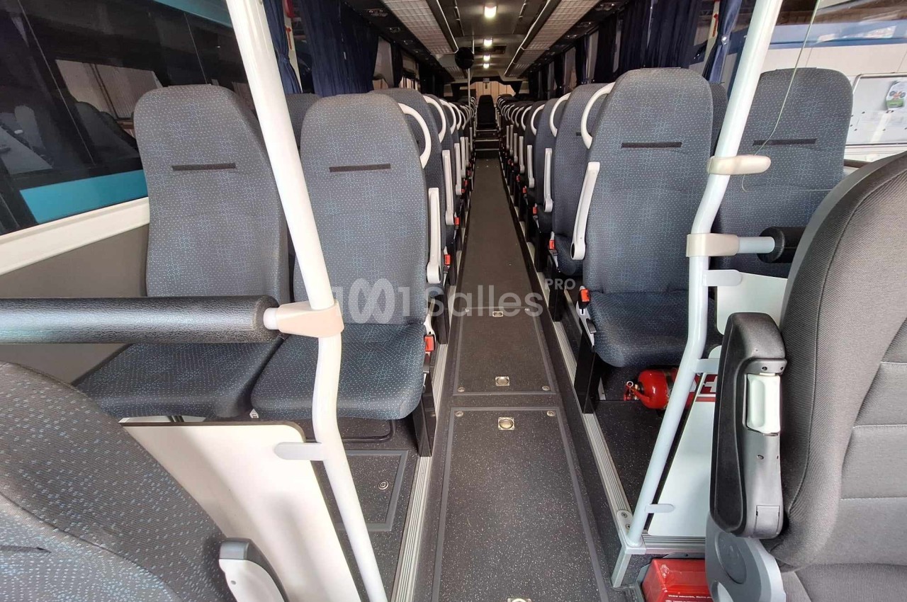 Intérieur d'un autocar avec rangées de sièges vides, allée centrale et barres de maintien visibles.