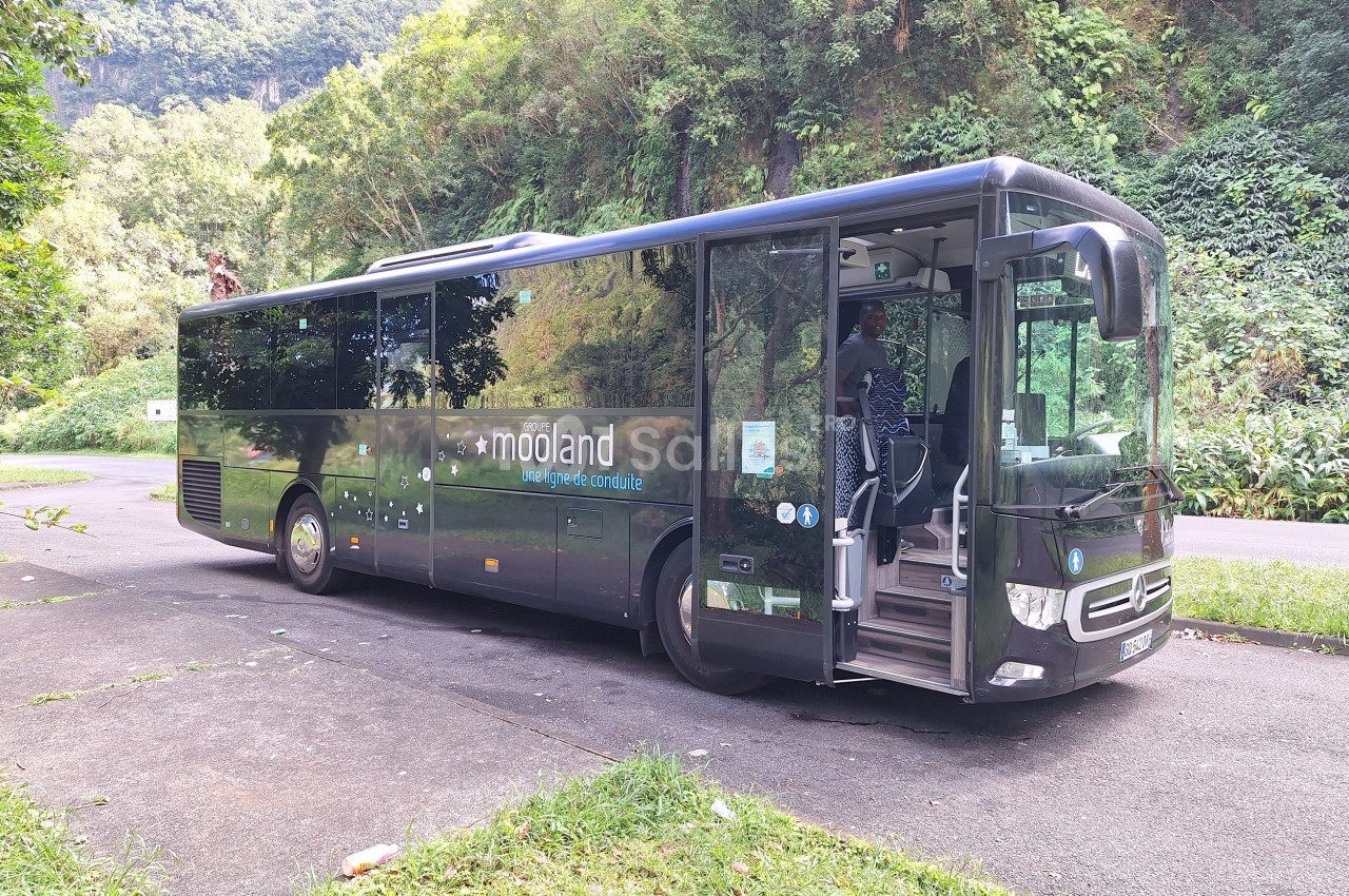 Autocar noir stationné sur une route entourée de végétation luxuriante, avec une personne montant à bord.