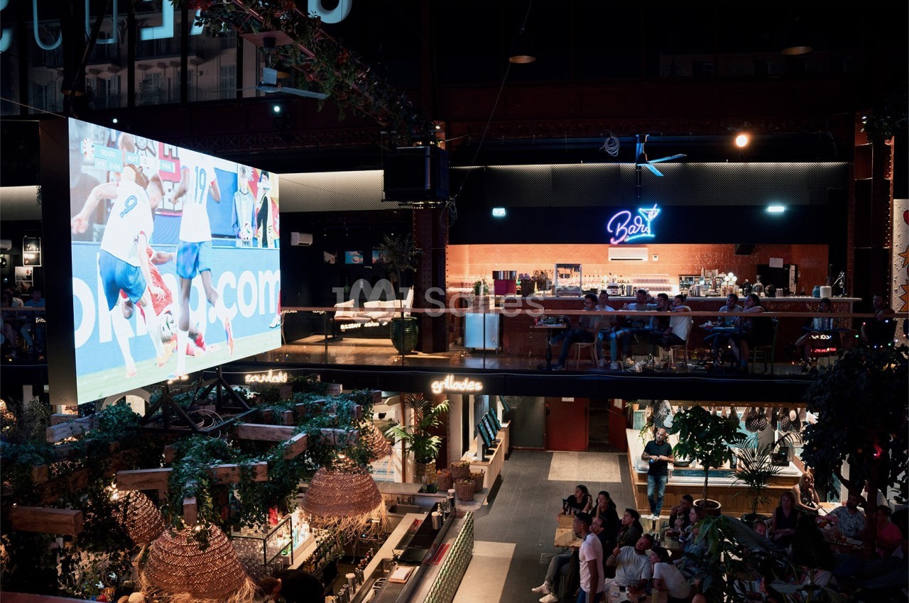 Grand écran diffusant un match de football dans un espace intérieur animé avec bar et spectateurs.