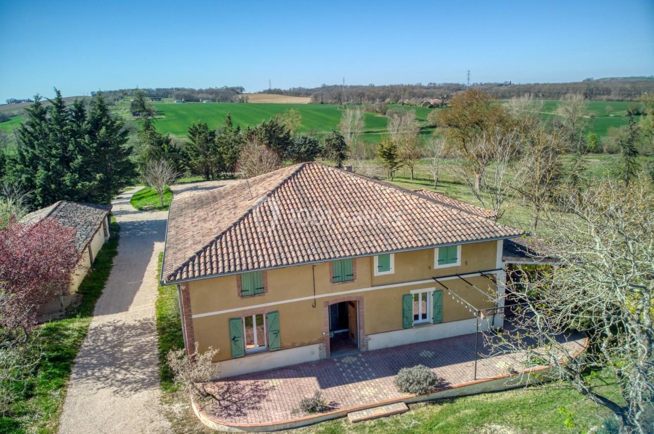 Maison à deux étages avec toit en tuiles, entourée de végétation et située dans un paysage rural verdoyant.