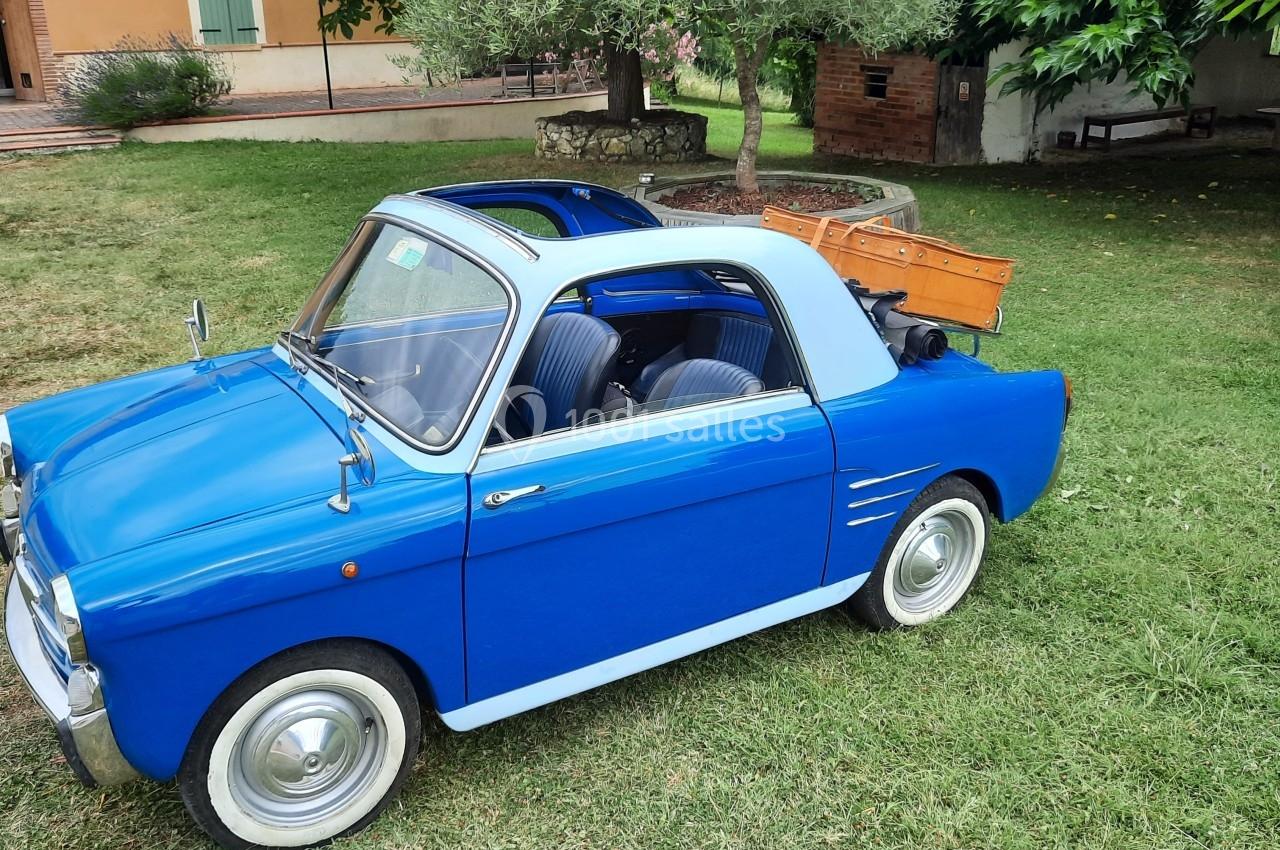 Petite voiture ancienne bleue avec toit ouvert, stationnée sur une pelouse près d'arbres et d'un bâtiment en arrière-plan.