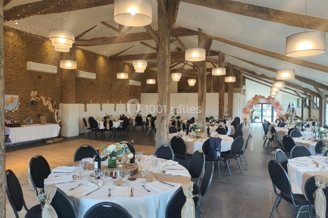 Salle de réception décorée pour un événement, avec tables dressées, chaises noires et éclairage suspendu.
