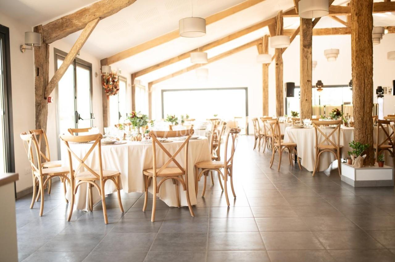Salle lumineuse avec tables rondes décorées, chaises en bois et poutres apparentes dans un cadre moderne et chaleureux.