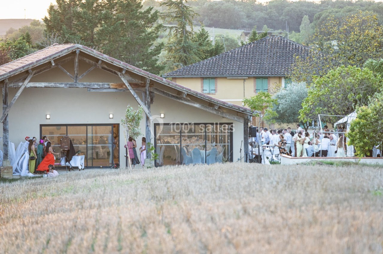Vue d'une réception en plein air près d'un bâtiment avec des invités rassemblés et un paysage champêtre en arrière-plan.