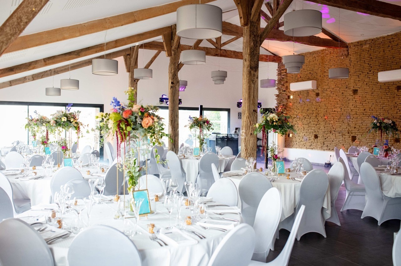 Salle de réception lumineuse avec tables rondes décorées de fleurs et chaises blanches, poutres apparentes au plafond.