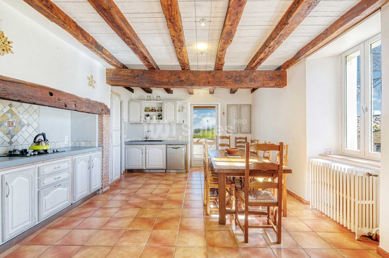 Cuisine lumineuse avec poutres apparentes, carrelage au sol, table en bois et vue sur l'extérieur par une porte vitrée.