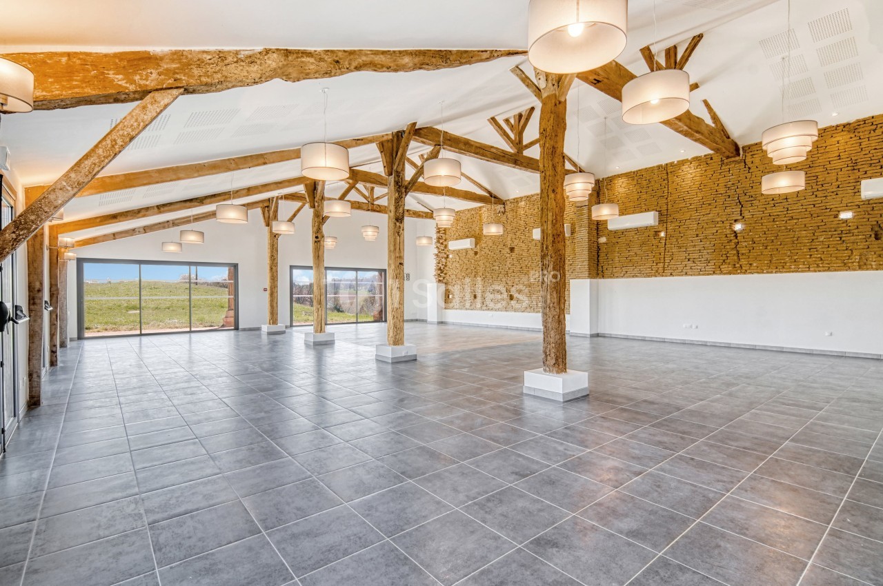 Grande salle lumineuse avec poutres apparentes, murs en briques et carrelage gris, donnant sur un espace extérieur.