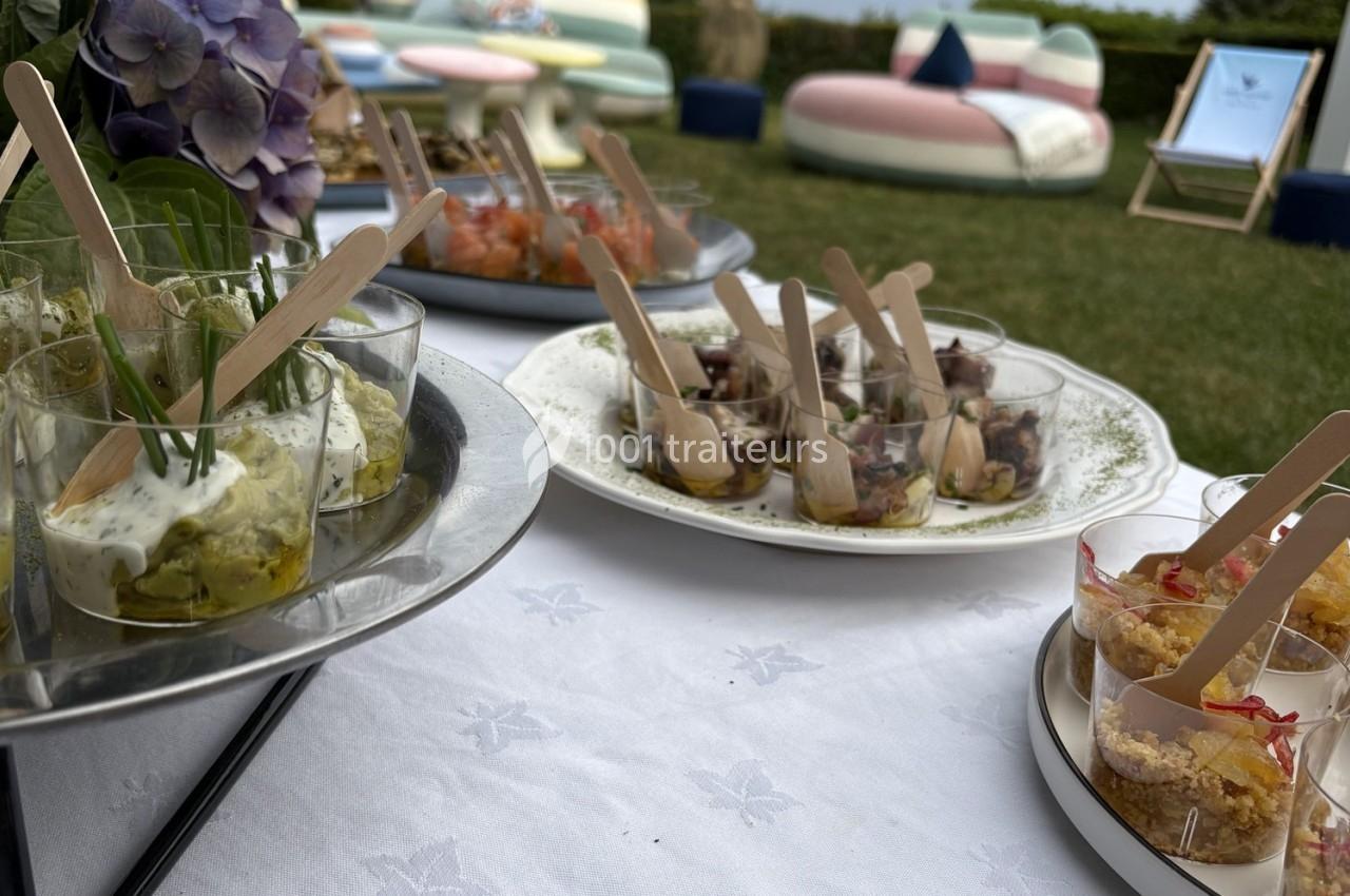 Plateaux de verrines variées présentées sur une table blanche en extérieur, avec des chaises et plantes en arrière-plan.