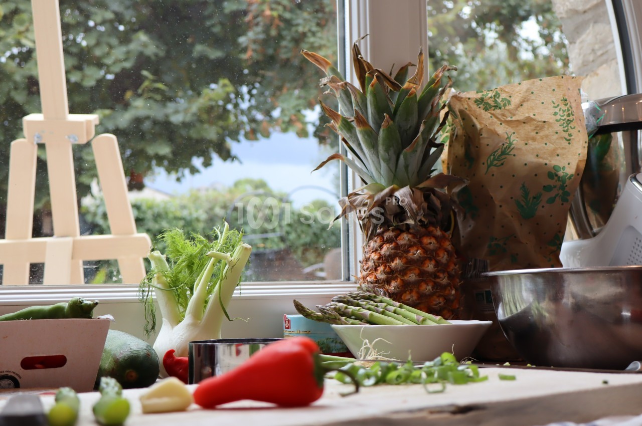 Fruits, légumes et ustensiles de cuisine disposés sur un plan de travail près d'une fenêtre donnant sur un jardin.