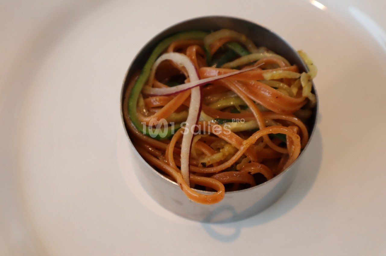 Assiette contenant des spaghettis colorés avec des légumes, présentés dans un cercle en métal.