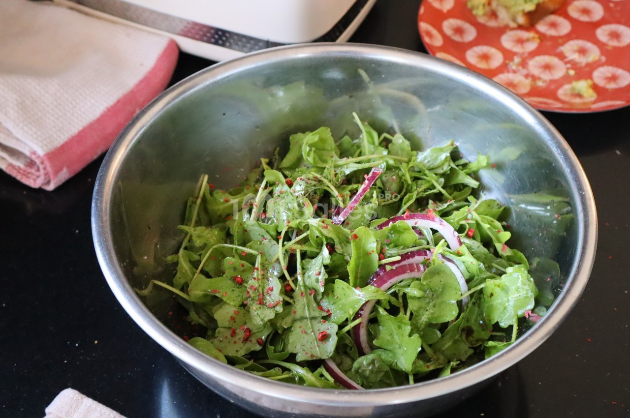 Salade de roquette fraîche avec rondelles d'oignon rouge et épices dans un saladier en métal sur un plan de travail.