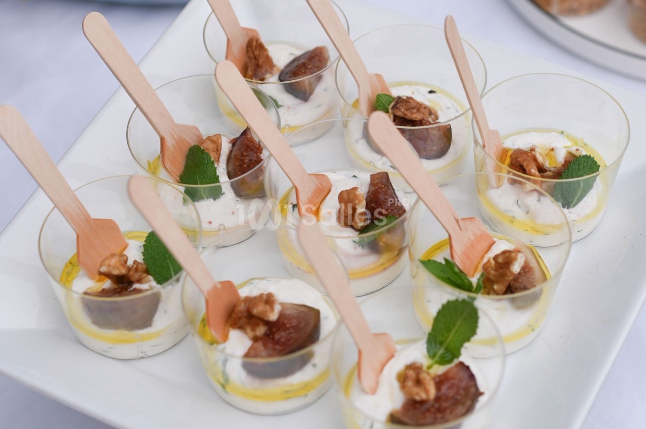 Verrines de fromage frais garnies de figues, noix, feuilles de menthe et cuillères en bois sur un plateau blanc.