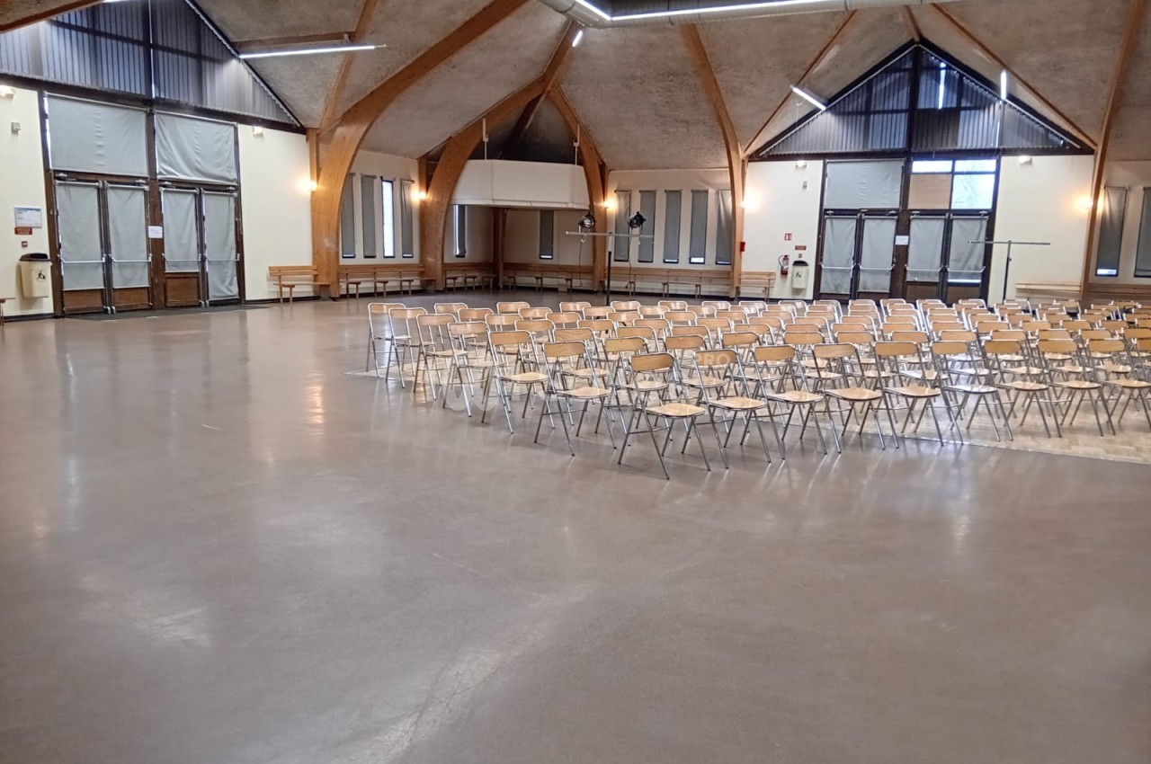 Salle spacieuse avec des rangées de chaises vides, éclairée par des fenêtres et des luminaires suspendus.