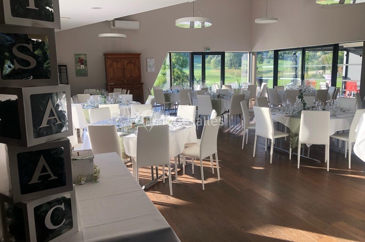 Salle de réception lumineuse avec des tables rondes dressées pour un événement, entourées de chaises blanches.