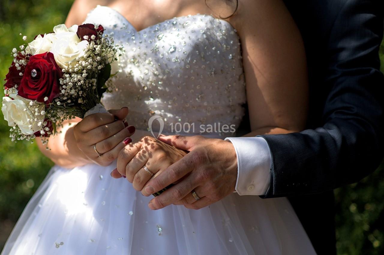 Un couple en tenue de mariage, tenant un bouquet de fleurs rouges et blanches, mains entrelacées.