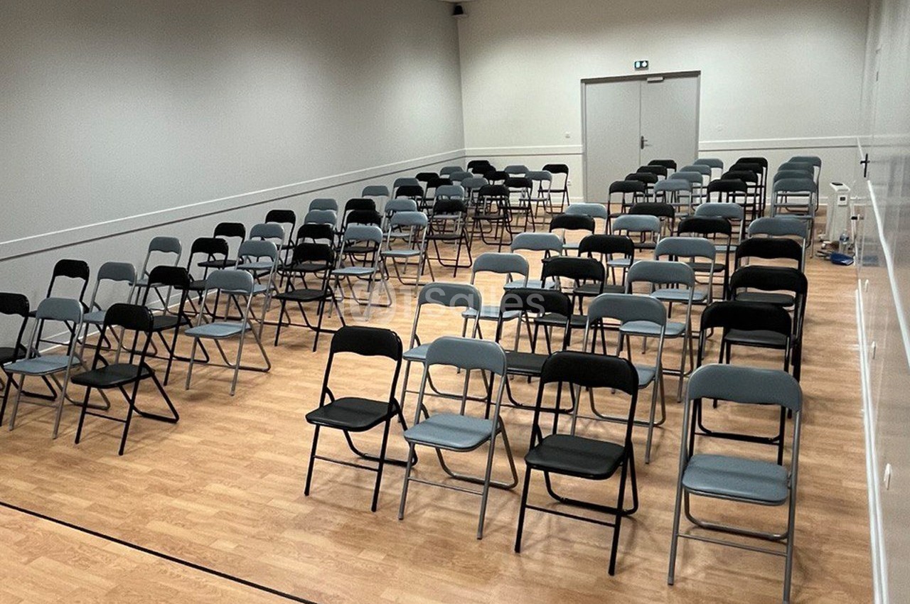 Salle avec des rangées de chaises pliantes noires et grises disposées sur un sol en bois clair, éclairée par des néons.