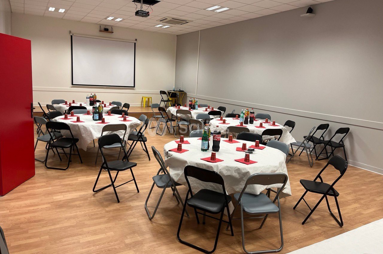 Salle aménagée avec des tables rondes dressées, nappes blanches, chaises pliantes noires et écran de projection en fond.
