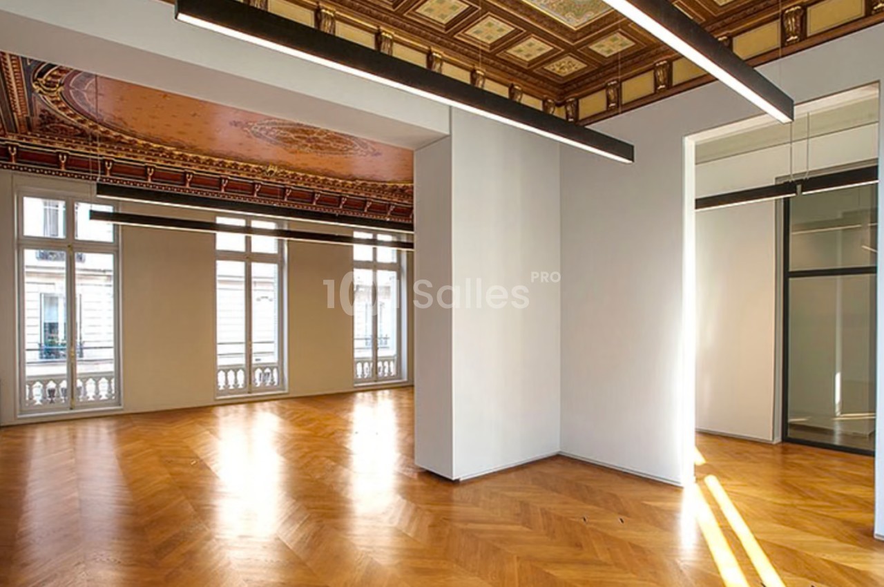 Salle lumineuse avec parquet en bois, plafond décoré et grandes fenêtres donnant sur l'extérieur.