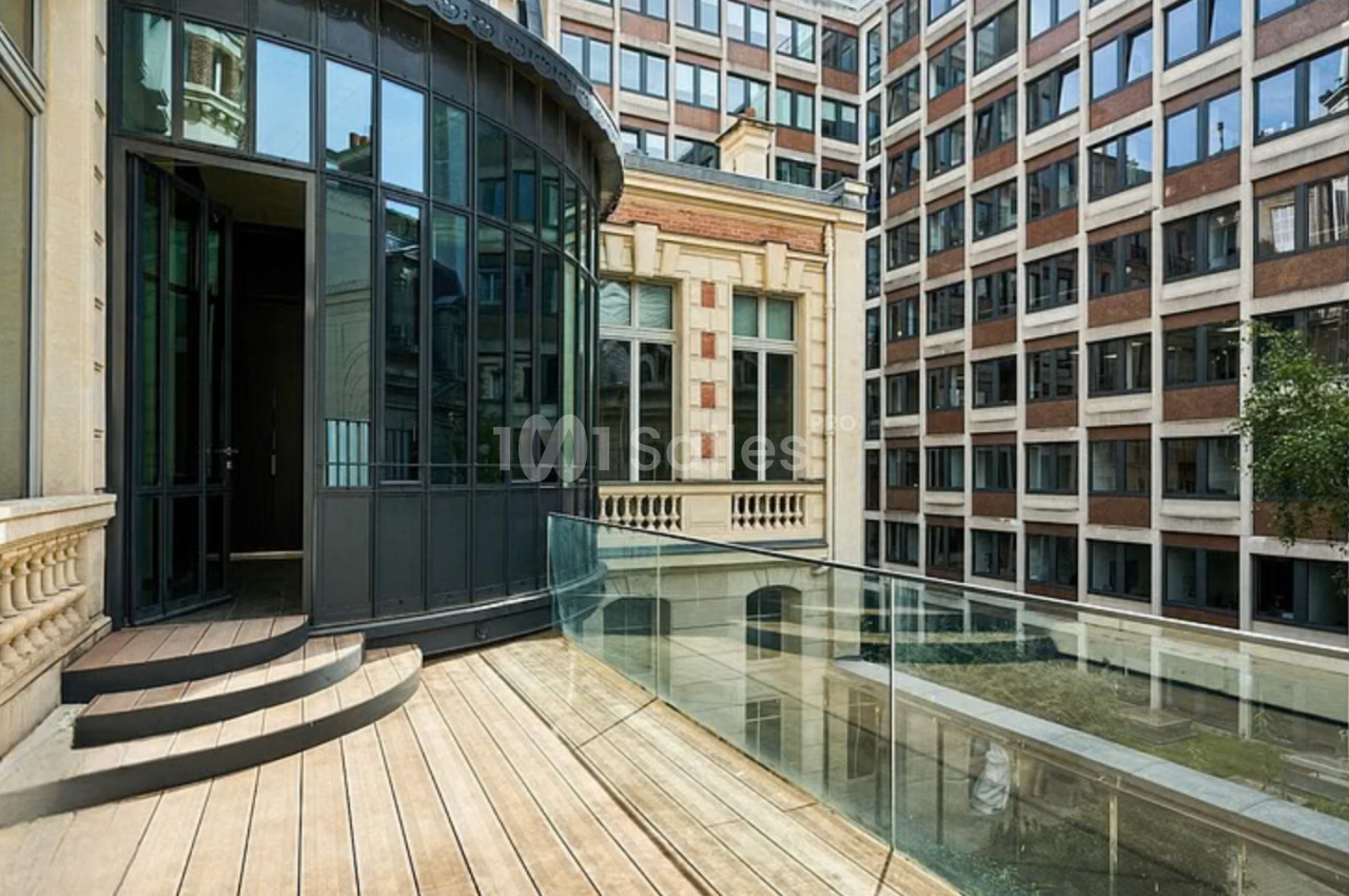 Terrasse en bois avec garde-corps en verre, donnant sur une cour intérieure entourée de bâtiments modernes.