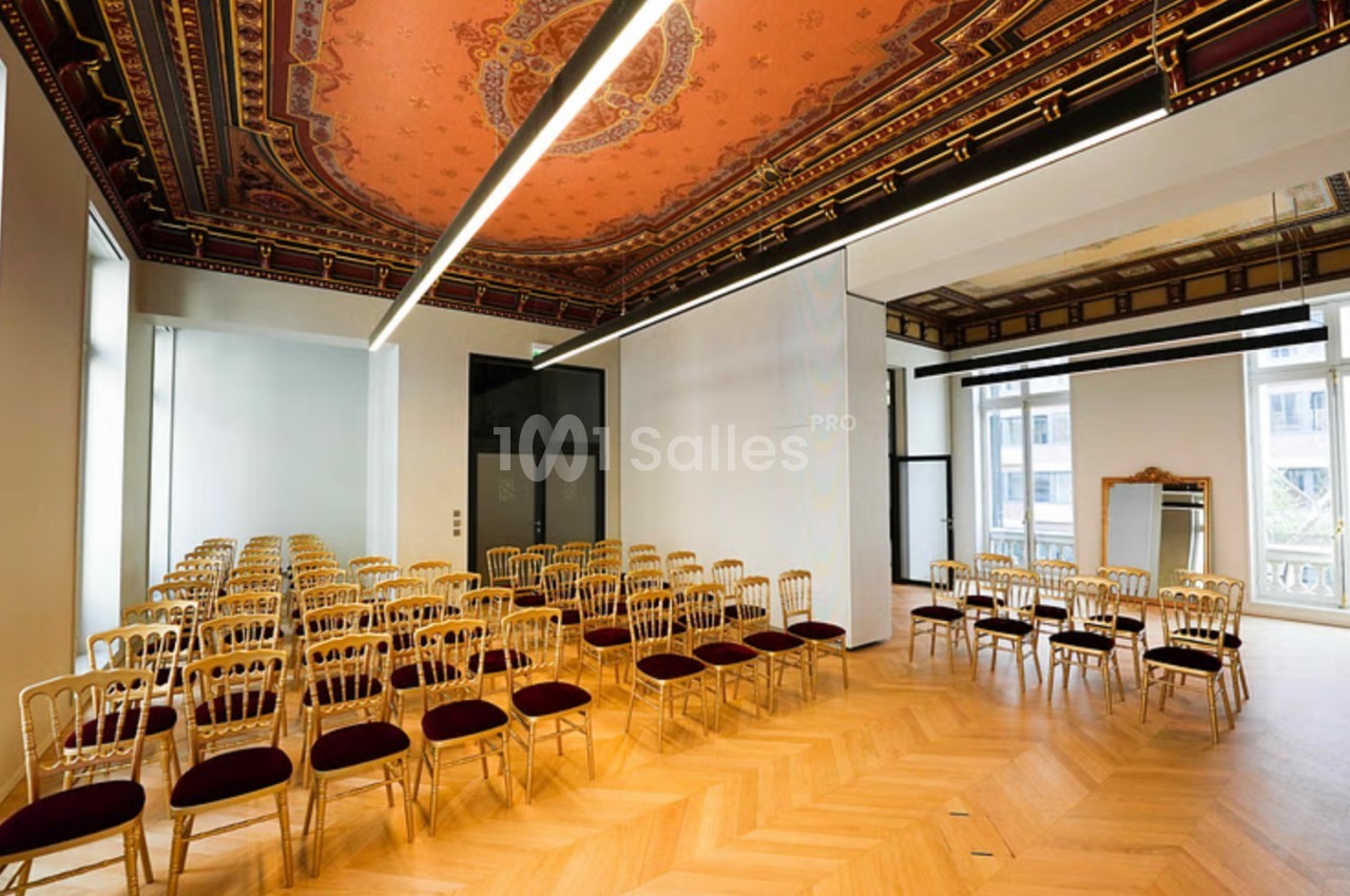 Salle lumineuse avec parquet en bois, plafond décoratif et chaises alignées pour un événement ou une réunion.