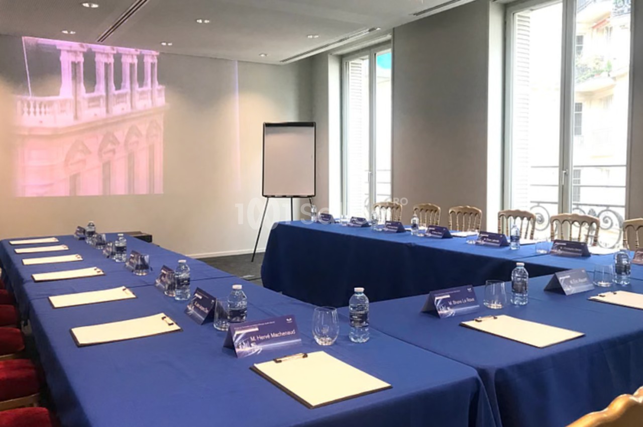 Salle de réunion avec tables couvertes de nappes bleues, chaises alignées, bloc-notes, bouteilles d'eau et un tableau blanc.