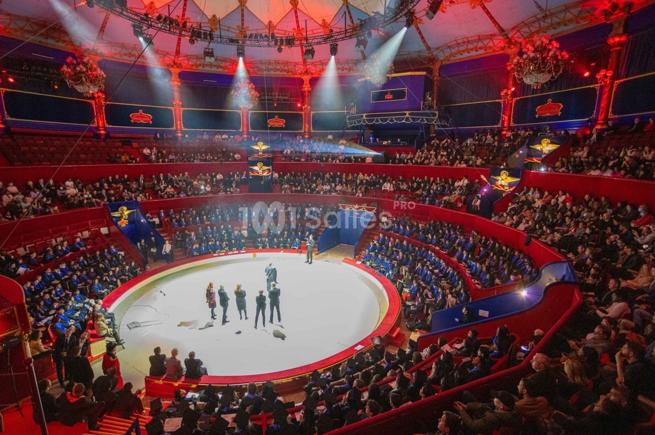 Vue d'une piste de cirque entourée de gradins remplis de spectateurs, éclairée par des projecteurs colorés.