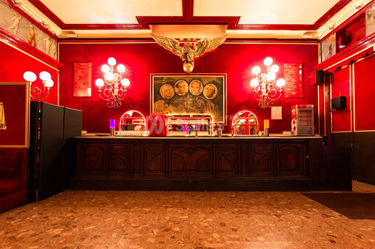 Comptoir en bois sombre dans un intérieur rouge avec lustres, tableau central et décoration dorée en forme d'aigle.