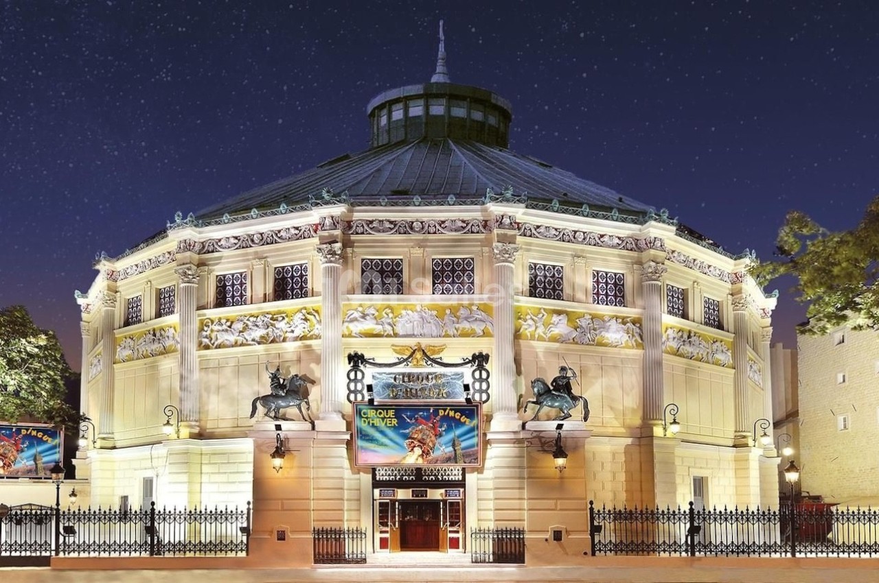 Façade illuminée d'un bâtiment circulaire avec des affiches de spectacle, vue de nuit sous un ciel étoilé.
