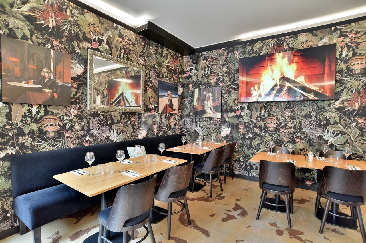 Salle de restaurant avec tables en bois, banquettes noires, murs décorés de papier peint tropical et cheminée virtuelle.