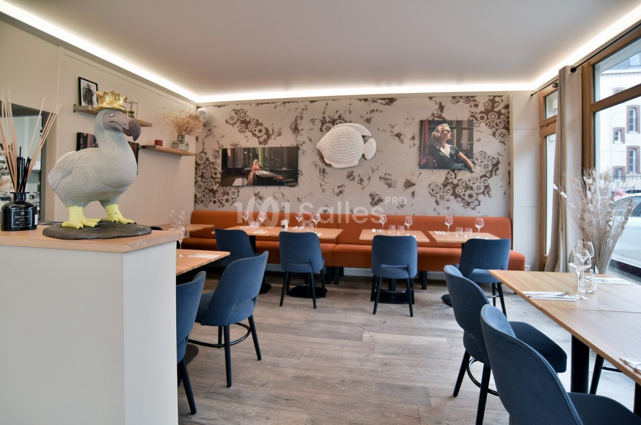 Salle de restaurant lumineuse avec tables en bois, chaises bleues, banquette orange et décoration murale artistique.